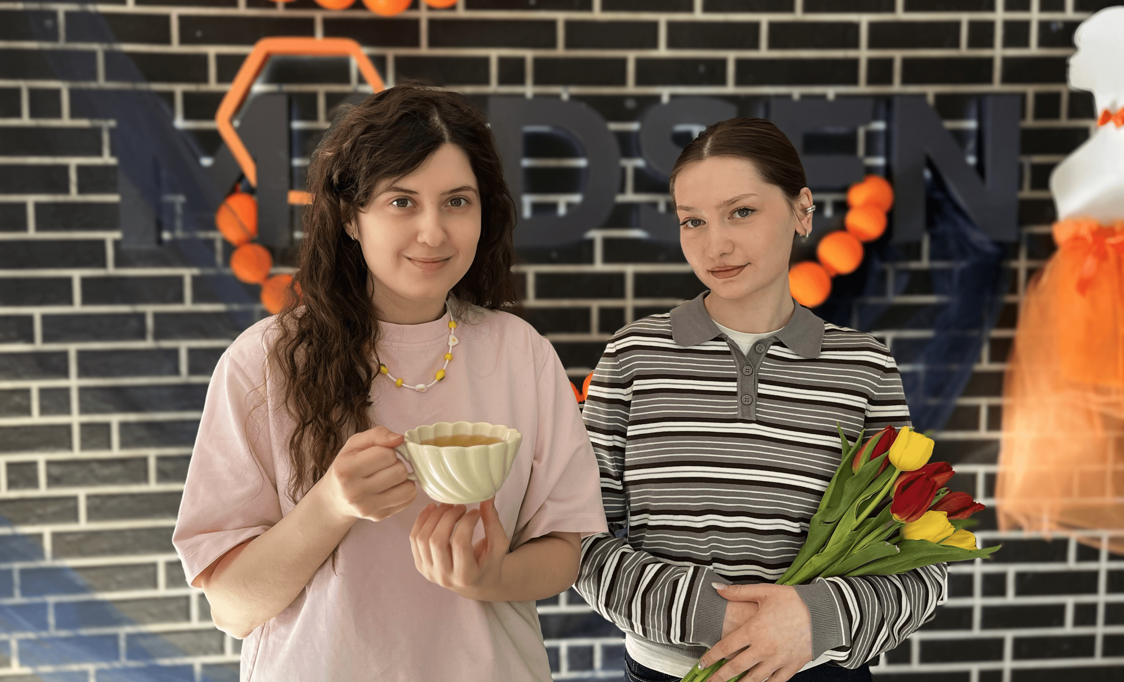 Two girls standing near Modsen International Women's Day decorations, holding flowers and a cup of tea Two girls standing near Modsen International Women's Day decorations, holding flowers and a cup of tea